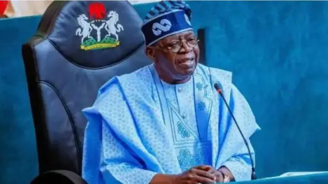 Aarẹ Bola Tinubu. O wọ agbada alawọ aro, o de fila alawọ kan naa, o lo igo soju, o si kawọ rẹ mejeeji sori tabili bo ṣe n rẹrin-in muse ninu aworan yii.