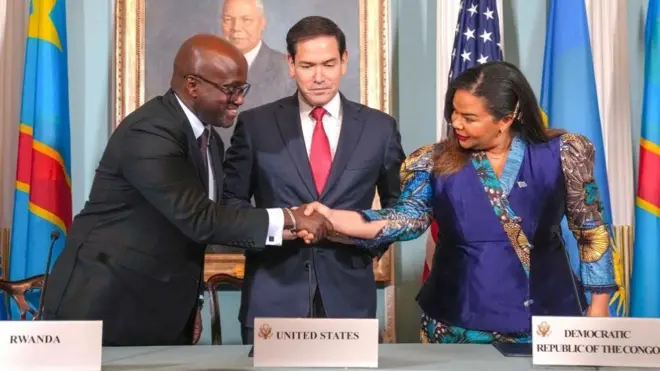 Minisitiri w'ububanyi n'amahanga w'u Rwanda Olivier Nduhungirehe asuhuzanya na Minisitiri w'ububanyi n'amahanga wa DRC Thérèse Kayikwamba. Bombi barahagaze kandi bafite akanyamuneza ku maso, hagati yabo hahagaze Minisitiri w'ububanyi n'amahanga w'Amerika Marco Rubio. Inyuma ku rukuta hari ifoto y'umugabo iri muri 'cadre' ndetse inyuma hanagaragara ibendera rya DRC n'iry'Amerika. Ifoto yo ku itariki ya 27 Kamena (6) mu 2025, yafashwe ikuwe muri videwo y'umuhango urimo kuba. 