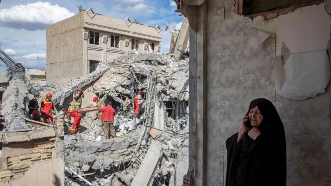 27 मार्च को तेहरान में हवाई हमले में क्षतिग्रस्त हुई एक रिहायशी इमारत के मलबा हटाते इमरजेंसी वर्कर्स और पास में फोन पर बात करती एक महिला. 