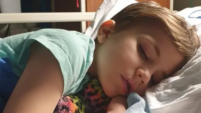 Roo, uma criança pequena deitada em uma cama de hospital, segurando um brinquedo