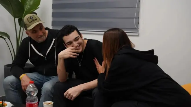 Edan Alexander sonriente en medio de sus dos padres, quienes lo miran con emoción