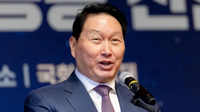 Close-up photo of Chey Tae-won speaking on a microphone wearing a suit and tie against a blurred blue and white background