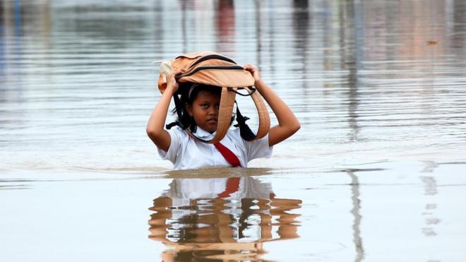 Una niña camina con su mochila sobre la cabeza y rodeada de agua hasta el pecho.