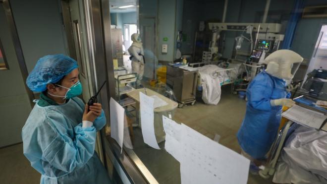 Três profissionais de saúde altamente protegidos com jalecos, máscaras e toucas trabalham em sala de internação em hospital de Wuhan; dois deles se falam através de aparelhos de rádio