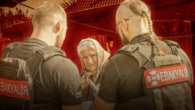 Двоє чоловіків у бронежилетах із написом «ЕВАКУАЦІЯ» спілкуються з літньою жінкою, яка тримає документи та стривожено дивиться повз камеру. Сцена виглядає як допомога цивільним у зоні небезпеки. 