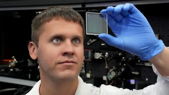 Ilya Kazansky, diretor executivo da SPhotonix, com roupa branca de laboratório e luvas de borracha azuis, observa uma pequena placa de vidro quadrada com marcas quadriculadas.