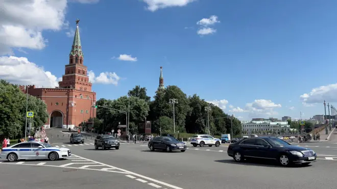 Вид на башню московского Кремля, от которой отъезжает кортеж из черных машин и двух полицейских автомобилей.