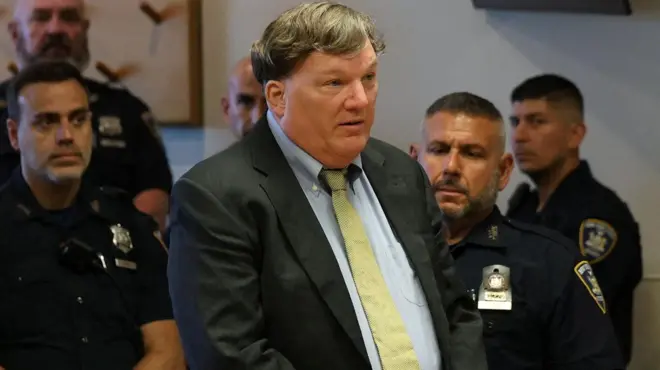 Rex Heuermann, con traje oscuro y camisa y corbata claras en el tribunal, con las manos entrelazadas y gesto serio, rodeado de agentes de policía y una mujer sentada a su derecha