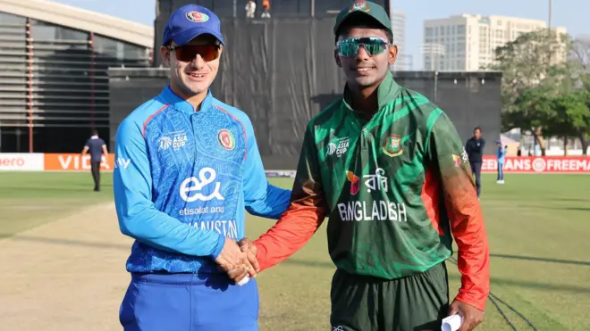 د افغانستان او بنګله دېش له نوولس کلنو د کم عمره لوبډلو لوبډلمشران چې د خپلو لوبډلو جامې یې پر تن او لمرینه ورځ یې کې د لمر تورې عینې اچولي خندانه څنګ په څنګ ولاړ دی او یو بله ته یې لاس ورکړی. دوی د ټاس له اچول کېدو وروسته عکس ته ولاړ دي او مخامخ کمرې ته ګوري.