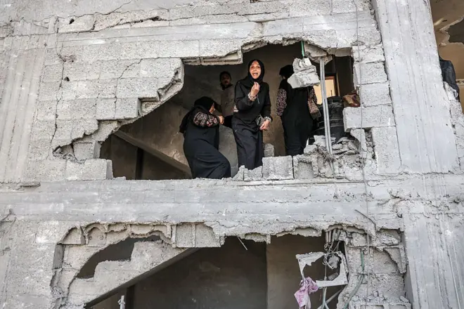 يظهر واضحاً في الصورة ثلاث نساء فلسطينيات يرتدين عباءات سوداء يقفن أمام فتحة في جدار مبنى مهدم في غزة. الصورة التقطت نهاراً.