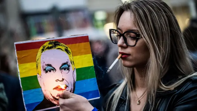 Devojka sa naočarima za sunce i cigarom u ustima u levoj ruci drži upaljač, a u desnoj poster sa Orbanovom slikom dok je pozadina zastava LGBT zajednice