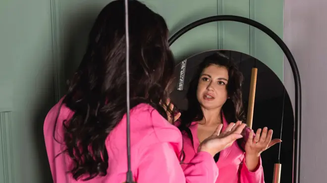 شابة تتدرب على إلقاء خطاب، وتتحدث إلى نفسها في المرآة، ترتدي قميصاً طويل الأكمام لونه وردي داكن.