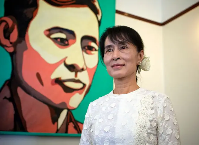 အစိမ်းရောင်နောက်ခံနဲ့ ဗိုလ်ချုပ်အောင်ဆန်းပုံပန်းချီကားရှေ့မှာ ရင်ဖုံးအင်္ကျီအဖြူ နှင်းဆီပန်းအဖြူပန်ပြီး မတ်တပ်ရပ်နေတဲ့ ဒေါ်အောင်ဆန်းစုကြည် 