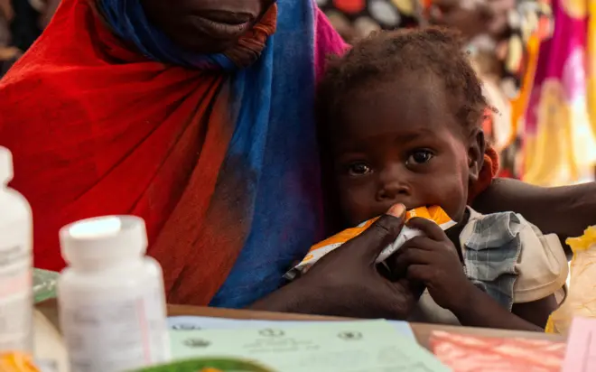 Sittna, two-years-old, eats her daily ration of MAM-treating supplements, while her mother Magedah gets instruction from the nutrition officer at the WFP-supported Philippe Nutrition Center, in Port Sudan, Red Sea State, Sudan May 2, 2024.