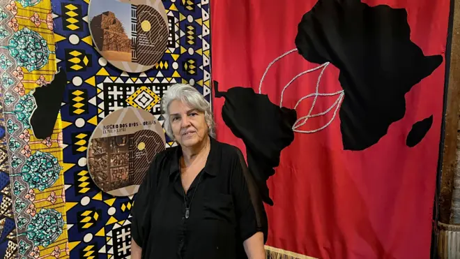 Uma mulher com cabelos grisalhos em pé. Ao fundo dela há um mural com um desenho do mapa da África ligado ao América do Sul