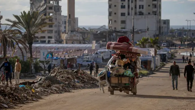 فلسطينيون يصلون إلى وسط مدينة غزة بعد نزوحهم من شمالي القطاع، يصلون مشياً أو في عربة قديمة. الصورة التقطت نهاراً قرب خيام ودمار حول الطريق.