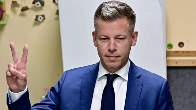 Mavi takım elbiseli kumral siyasetçi parmaklarıyla zafer işareti yapıyor.