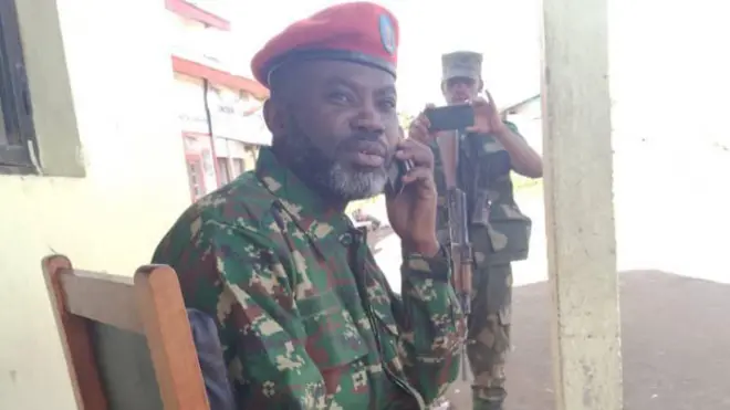 John Imani Nzenze (ku ifoto) yambaye imyenda ya gisirikare yiganjemo amabara y'icyatsi kibisi n'ikigina n'andi ajya kuba umweru. Yicaye ku ntebe ya rwego ikoze mu giti iriho umusego, arimo kuvugira kuri telefone. Yambaye ingofero itukura. Afite ubwanwa burimo imvi. Iruhande rwe hahagaze umugabo ufite imbunda urimo gufotoresha telefone. Yambaye ingofero n'imyenda ya gisirikare yijimye. Bombi barareba mu cyerekezo cy'ahari 'camera'. 