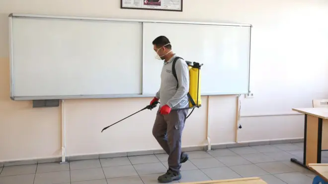 Boş bir sınıfı dezenfekte eden erkek görevli tahtanın önünde duruyor. 