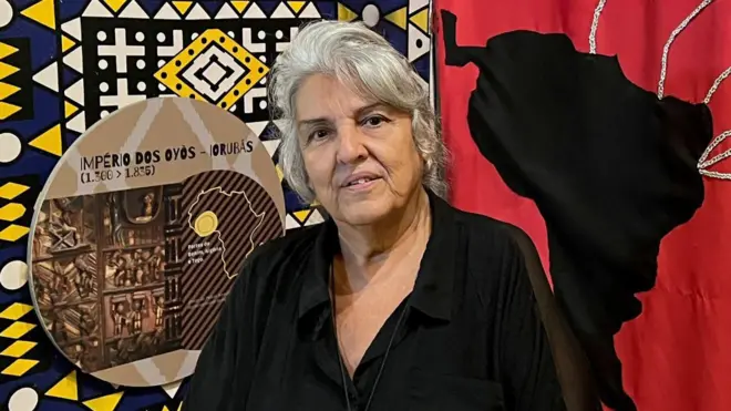 Una mujer de cabello gris, de pie. Detrás de ella hay un mural que muestra un dibujo del mapa de África conectado con América del Sur.
