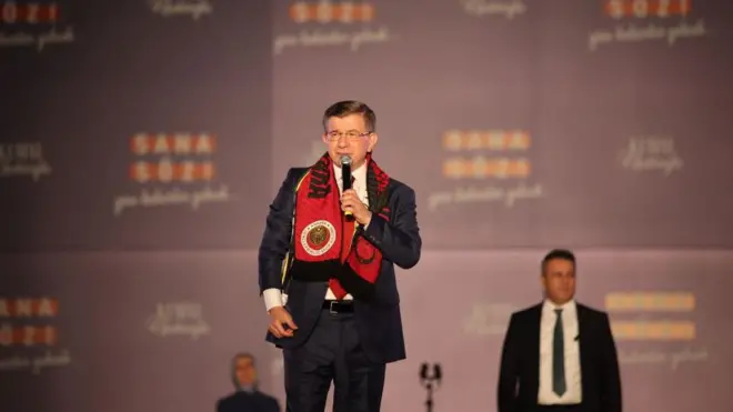 Davutoğlu kırmızı bir atkıyla bir miting sahesinde elinde mikrofonlar ayakta, resmi giyimli.