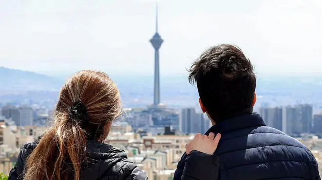 زن و مردی از بالای بلندی به نمایی از تهران که برج میلاد در آن مشخص است نگاه می کنند