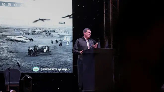 Abdi bir kürsüde konuşuyor arkasında duvara bir resim yansıtılmış, siyah beyaz bir şehri gösteriyor.