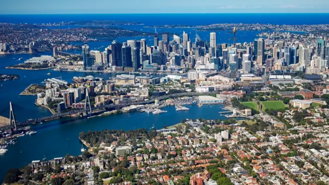 An aerial view of the Sydney city skyline with views of the harbour, rows of inner city houses and green spaces. Rows of orange roofs dot the foreground, with the blue water of the harbour running in between white and black skyscrapers and Sydney Tower in the background on sunny day.