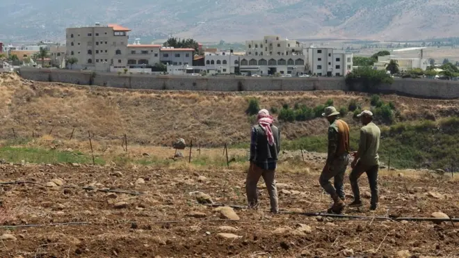 Three farmers walking in soil across a field in southern Lebanon, with a long wall and buildings in the background (file photo)