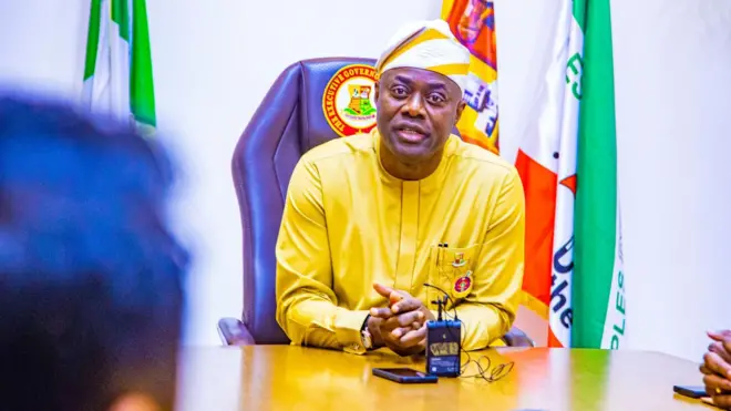 Gómìnà Seyi Makinde wọ aṣọ èsúrú, jòkòó lórí àga ó ká ọwọ́ sórí tábìlì, ó ń wo ọ̀ọ́kán