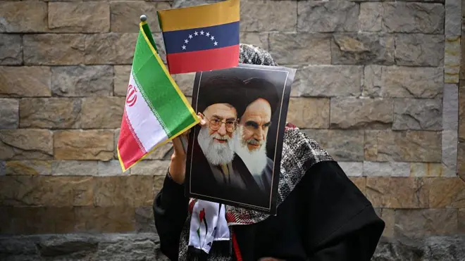 Tahran'da bir kadın elinde Humenyni ve Hamaney posterleri ile Venezuela ve İran bayrakları tutuyor. Tuttuğu şeyler yüzünü de kapatıyor