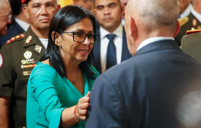 Delcy Rodriguez is wearing a green suit and glasses while greeting greets aa man whose back is toward the camera. sShe has her hand on his arm and is smiling. A man in uniform and a crow is behind her