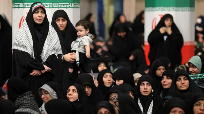 Siyah çarşaf giyinmiş İranlı kadınlar kapalı bir alandalar, arkalarındaki sütunlarda İran bayrakları giydirmesi var. Kadınları bazıları ayakta, çoğu yerde oturuyor. 