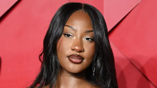 Tems regarde au-delà de la caméra, elle a de longs cheveux noirs et raides et se tient devant un fond rouge.