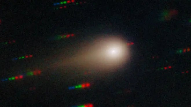 Комета 3I/Atlas показана в виде яркой точки, окруженной туманной сферой и хвостом слева. На заднем плане видны полоски синих, зеленых и красных точек, представляющие звезды, которые движутся относительно кометы.