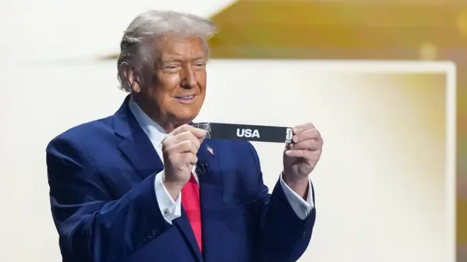 スーツに白シャツ、赤ネクタイ姿のトランプ氏が両手でくじをもって見せて笑顔を見せている