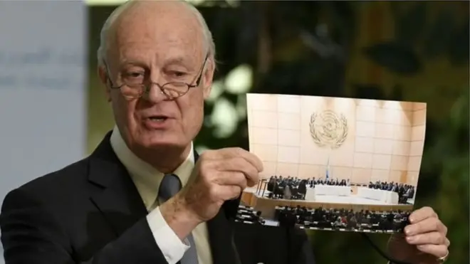 اقوامِ متحدہ کے شام میں سفیر سٹیفن ڈی مستورا شام کے متحارب گروہوں کی ملاقات کی تصویر دکھاتے ہوئے