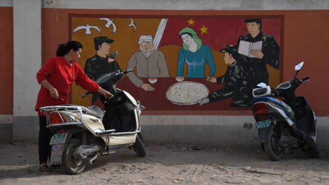 新疆喀什一位维吾尔族女士在一幅官方宣传民族政策壁画前停放摩托车（资料图片）