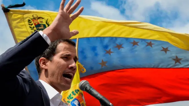 Venezuela National Assembly leader Juan Guaidó giving a speech at a microphone, with a Venezuelan fag fluttering behind him