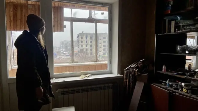 Анастасія у квартирі батьків, де родина не живе після того, як вибухова хвиля пошкодила будинок. З балкону видно розбомблений будинок, від вибуху в якому постраждав дім Анастасії.