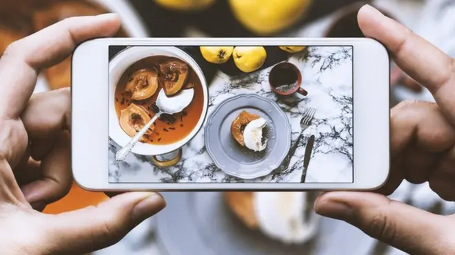 Hand taking top view photo of marble countertop. Cooking pot with poached quinces, poached quince with cream, fresh quinces and a cup of coffee on the countertop.