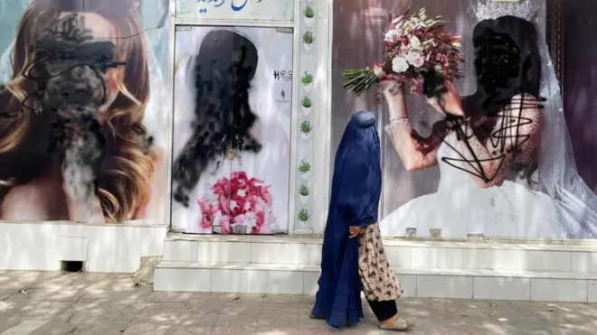 Mujer afgana camina frente a vallas publicitarias que muestran retratos de mujeres vandalizados.