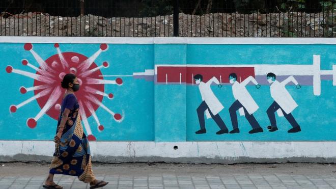 A woman wearing a mask walking past graffiti of three doctors injecting an virus