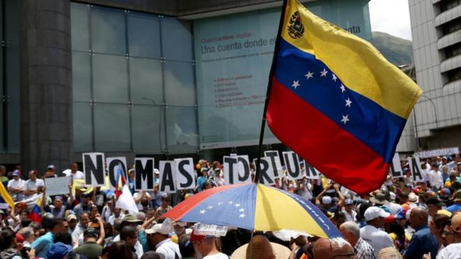 Bandera de Venezuela y detrás unos carteles que dicen "No más torturas".