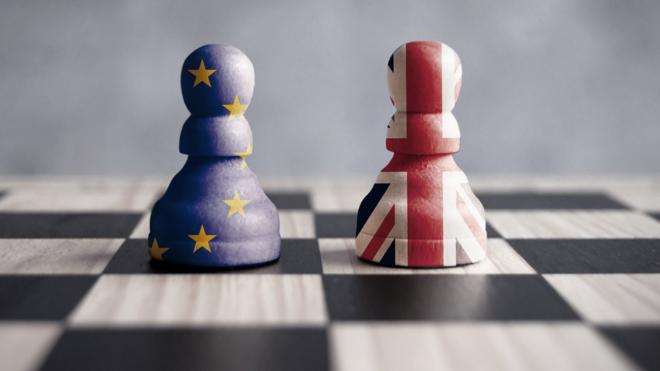 Two chess pieces on a chess board, one enclosed in the EU flag, one enclosed in the union jack.