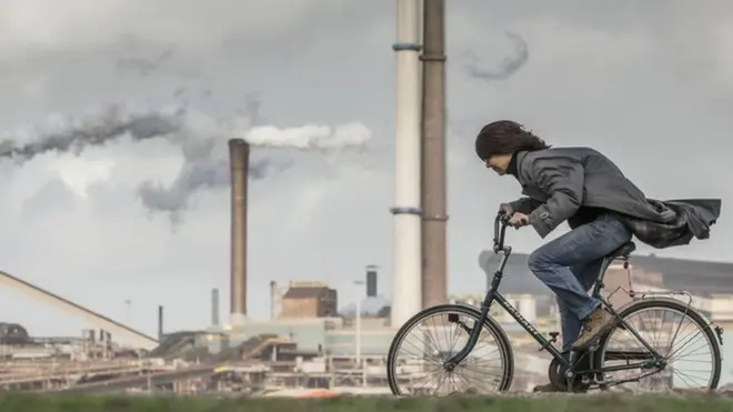Tata Steel factory, blast furnaces. Cyclist, woman, tries to stay upright in storm