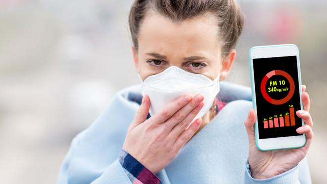 Mujer con una máscara y un celular que muestra el nivel de partículas PM10