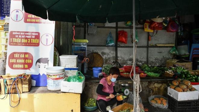Một người bán trái cây ở Hà Nội bên cạnh biển hiệu kêu gọi mọi người đeo khẩu trang để phòng ngừa và hạn chế sự lây lan của Covid-19