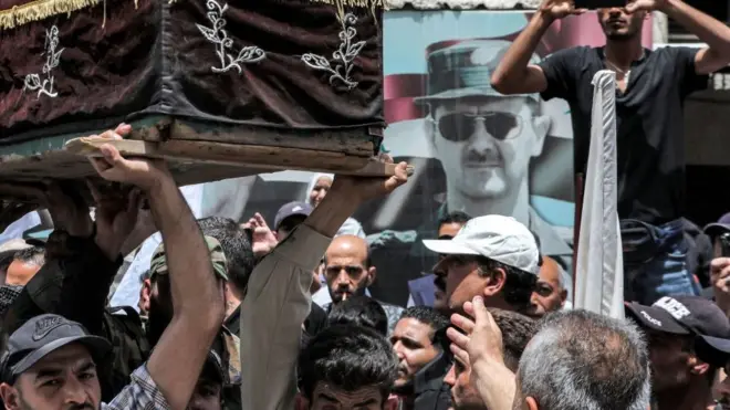 صورة للرئيس السوري بشار الأسد تلوح في الأفق على الحائط بينما يحمل المشيعون نعوش القتلى من أعضاء الجبهة الشعبية لتحرير فلسطين - القيادة العامة خلال جنازتهم بعد عودتهم إلى وطنهم في مخيم اليرموك للاجئين الفلسطينيين جنوب دمشق في 1 يونيو/ حزيران 2023.