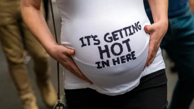 임신한 여성이 입고 있는 티셔츠에 "이곳은 더워지고 있다"고 쓰여 있고 여성이 글씨가 쓰여진 부분 배를 잡고 있다